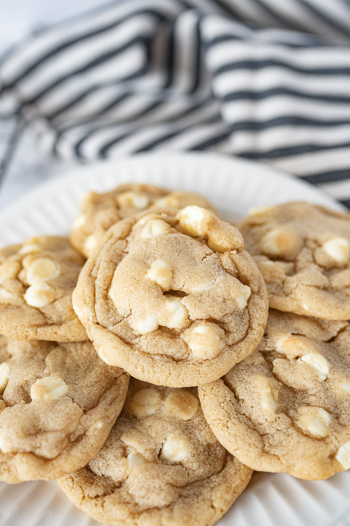 A pile of white chocolate chip cookies on a white plate with a striped towel in the background.