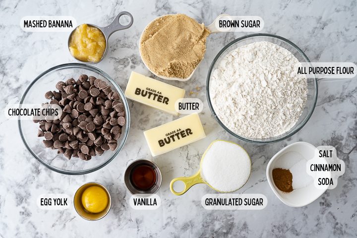 Ingredients for cookies on the counter including mashed banana, flour, chocolate chips, butter, an egg yolk, sugar, brown sugar, and cinnamon.