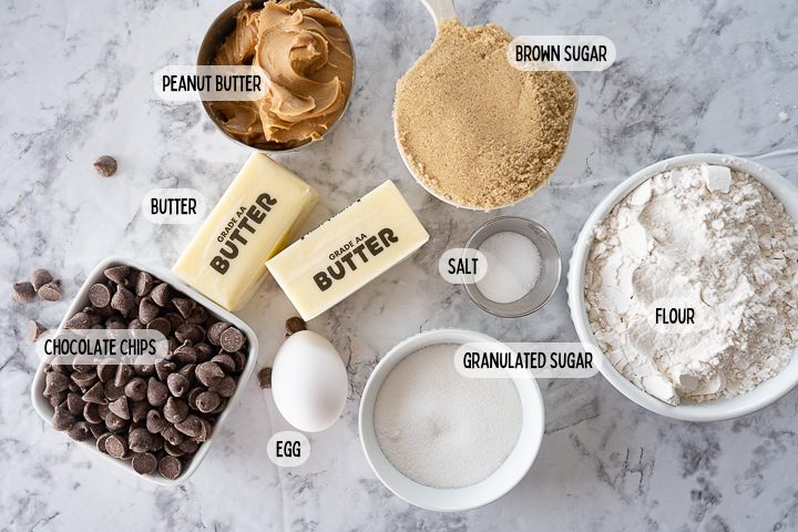 Ingredients in small dishes on the counter including peanut butter, butter, brown sugar, chocolate chips, an egg, granulated sugar, salt, and flour.