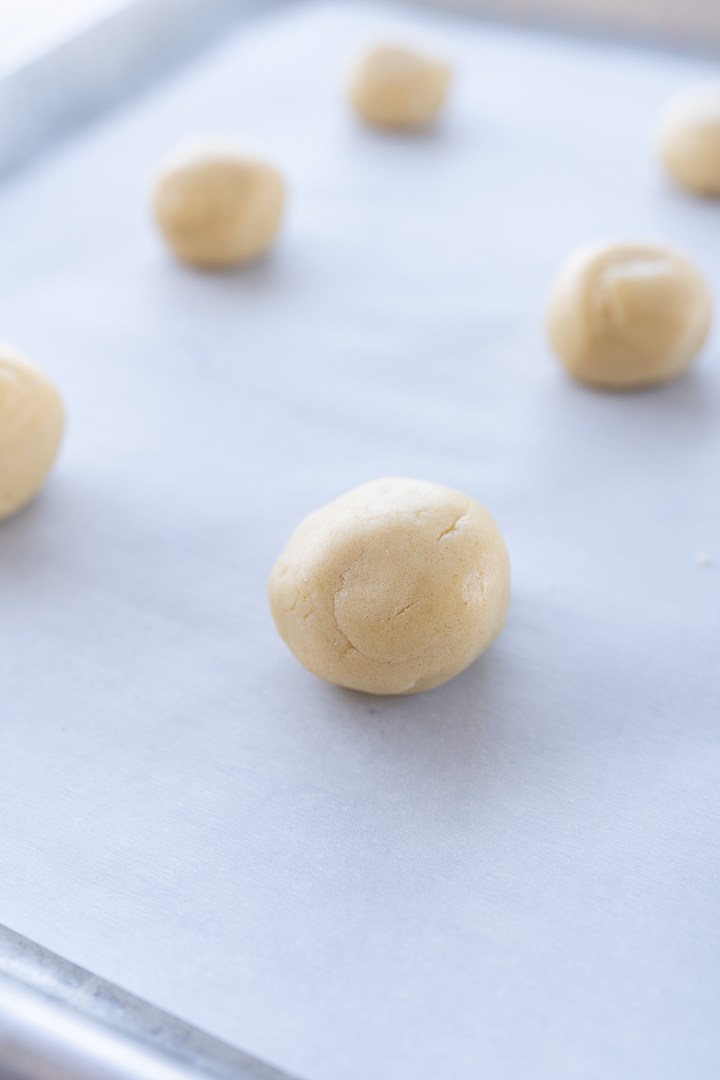 Six balls of sugar cookie dough on parchment paper on a baking sheet.