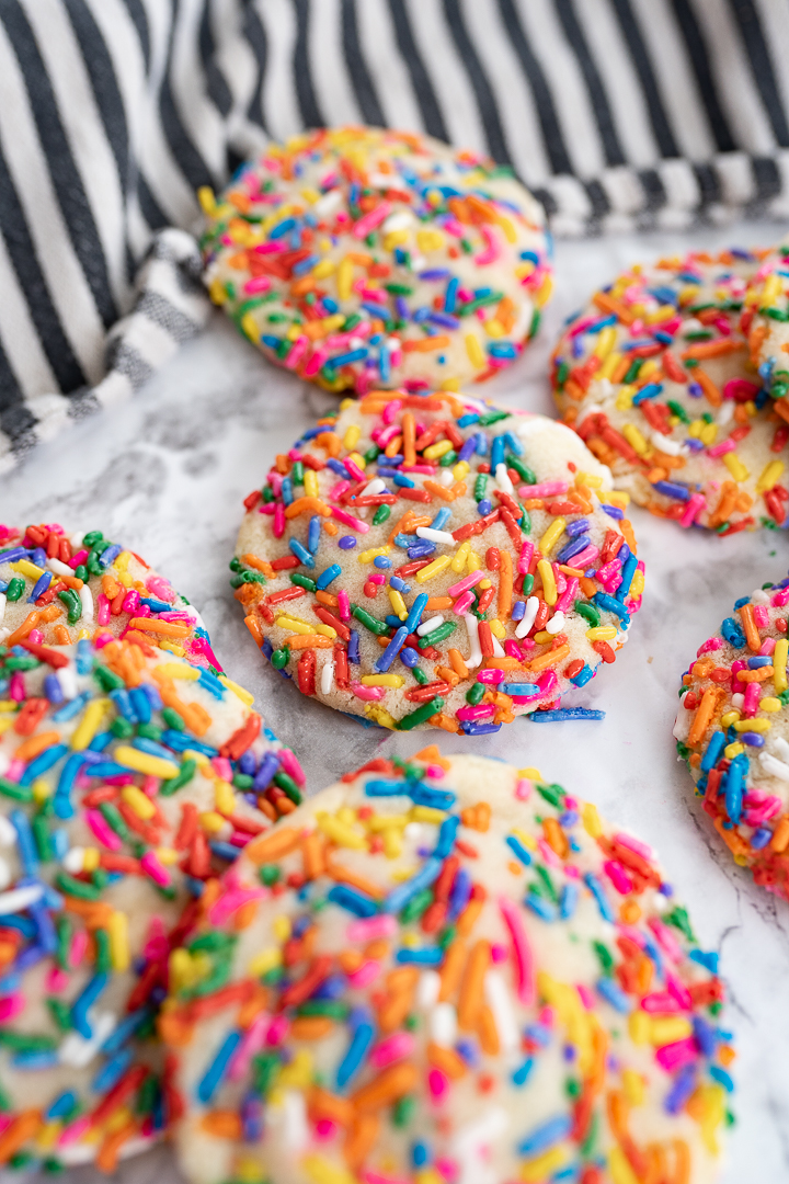 Several rainbow sprinkle cookies on the counter with a striped dish towel in the background.