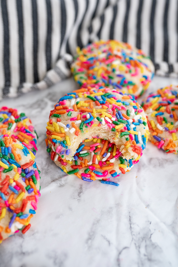 Several sprinkle cookies on the counter with a striped towel in the background and a bite taken out of one cookie.