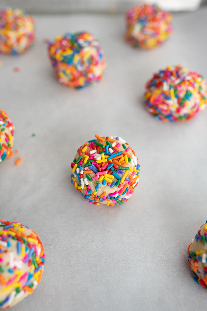 Sugar cookie dough balls rolled in rainbow sprinkles, sitting on parchment paper before baking.