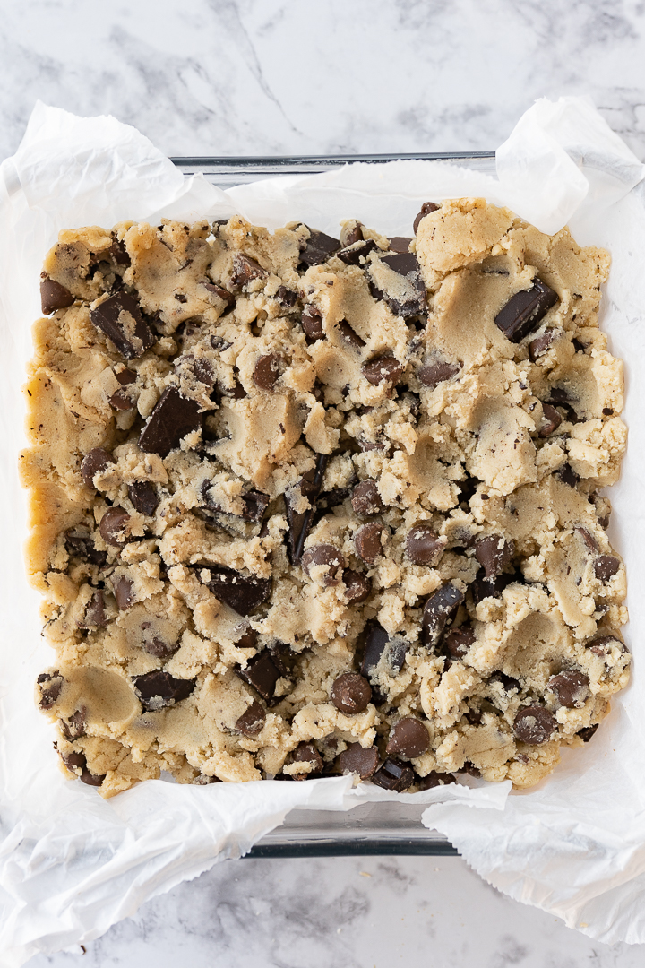 Chocolate chip cookie dough pressed into a baking pan with parchment paper on the counter.