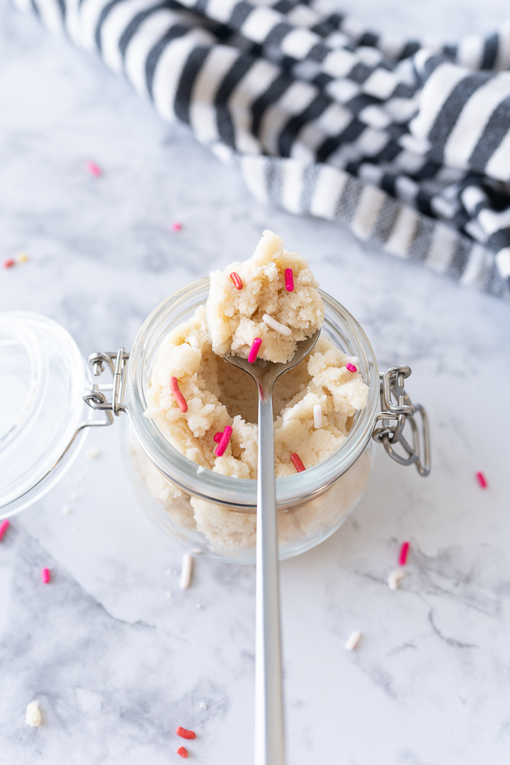 A jar with cookie dough and pink sprinkles with a spoon scooping out a bite.