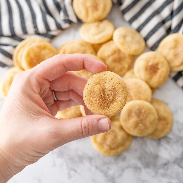 A hand holding a mini snickerdoodle cookie with several more snickerdoodles on the counter in the background.