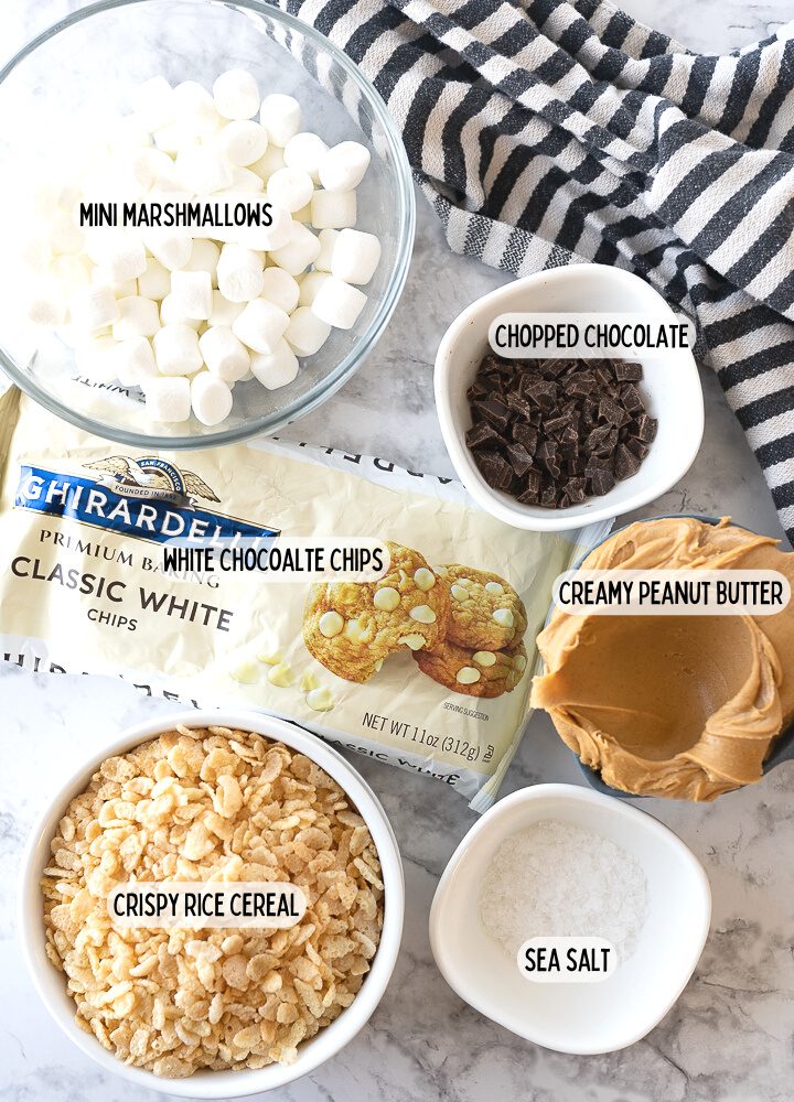 Ingredients in small bowls on the counter including marshmallows, chopped chocolate, peanut butter, crispy rice cereal, white chocolate chips, and salt.
