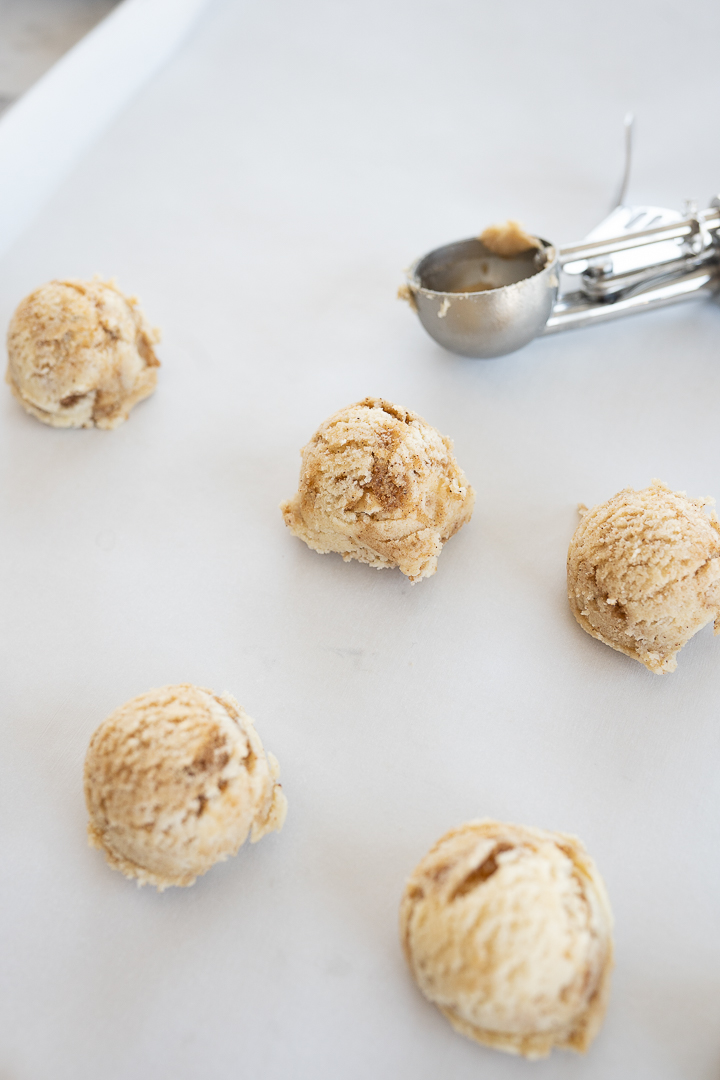 Balls of cinnamon roll cookie dough on parchment paper with a cookie scoop in the background.
