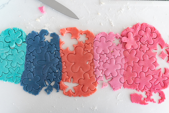 Light blue, dark blue, orange, light pink, and dark pink sections of sugar cookie dough rolled out on the counter with several flower shapes cut out of each section of dough.