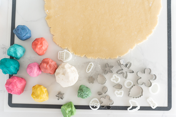 A large circle of plain cookie dough rolled out on the counter, with several different colored balls of cookie dough on the side, as well as various mini cookie cutters.