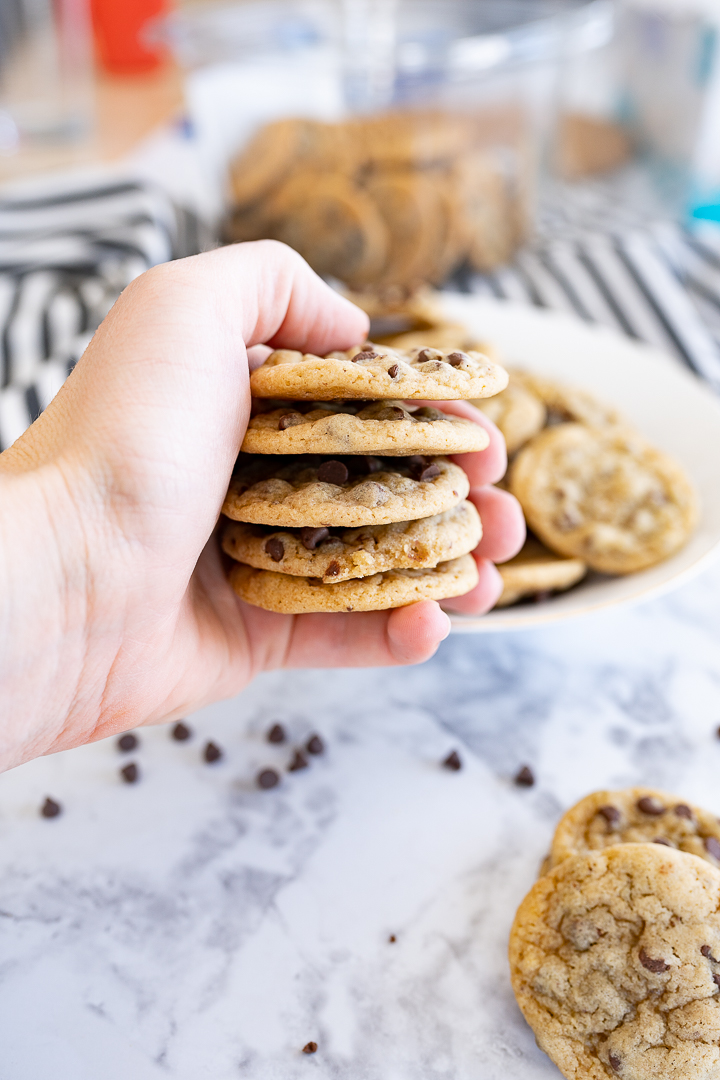 A hand holding a stack of mini chocolate chip cookies with more cookies and chocolate chips in the background.