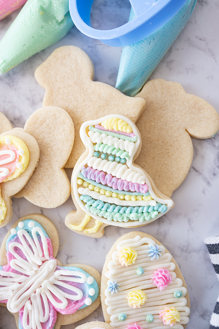 Up close photo of a chick shaped sugar cookie on the counter frosted with pastel rainbow frosting and piping bags in the background.