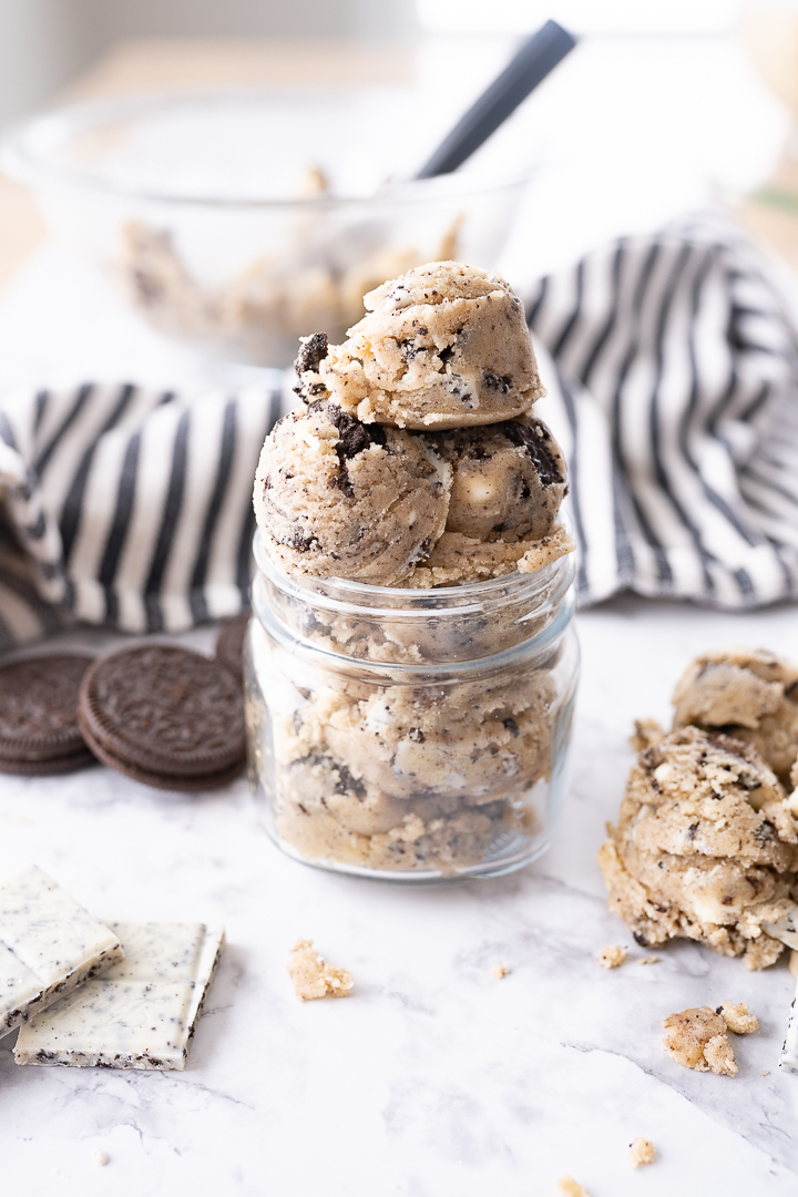 A glass jar of edible cookie dough on the counter with oreos and a striped dish towel in the background.
