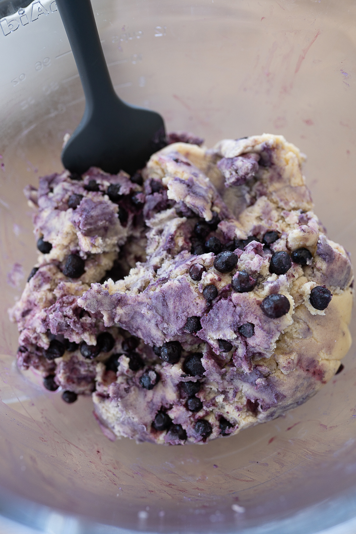 Purple and tan Lemon Blueberry cookie dough in a bowl. Blueberries are being folded in with a spatula. 