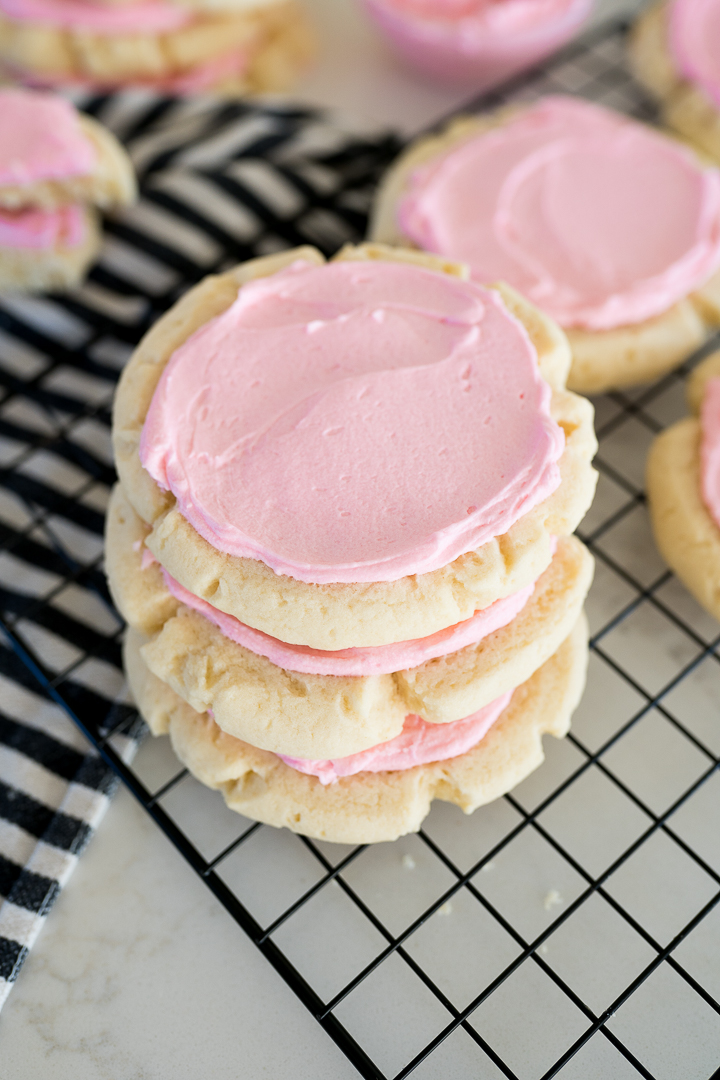 Crumbl sugar cookies with pink frosting stacked on a wire cooling rack 