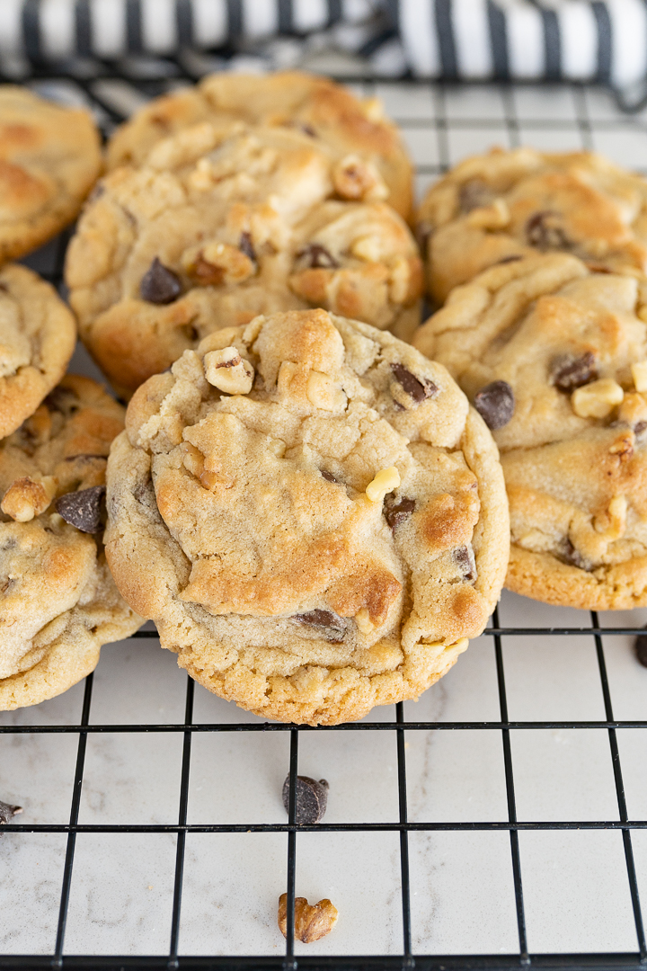 A pile of cookies on a wire cooling rack with chocolate chips and walnuts falling off onto the counter.