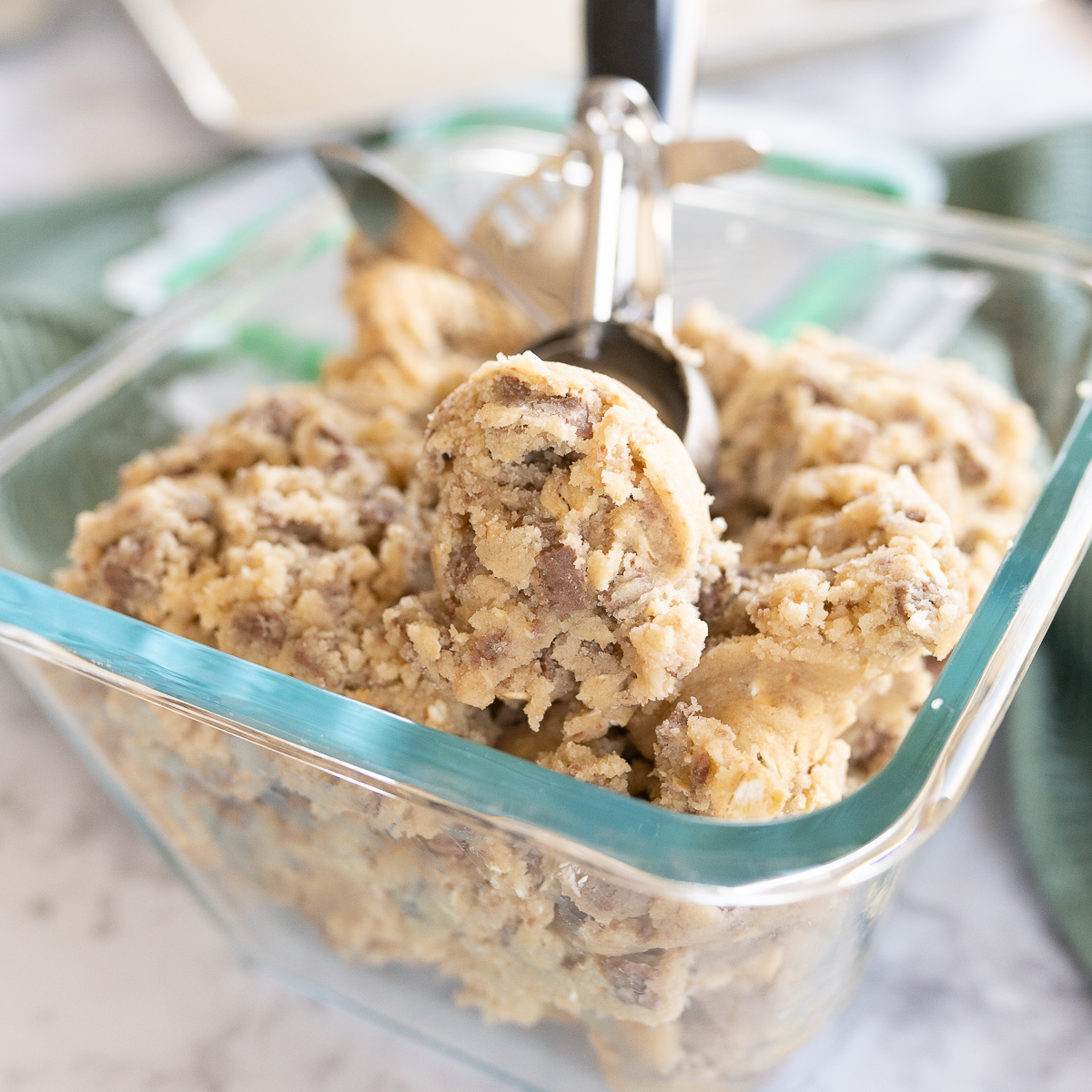 Cookie dough in a glass container without the lid. A cookie scoop is placed in the dough. 