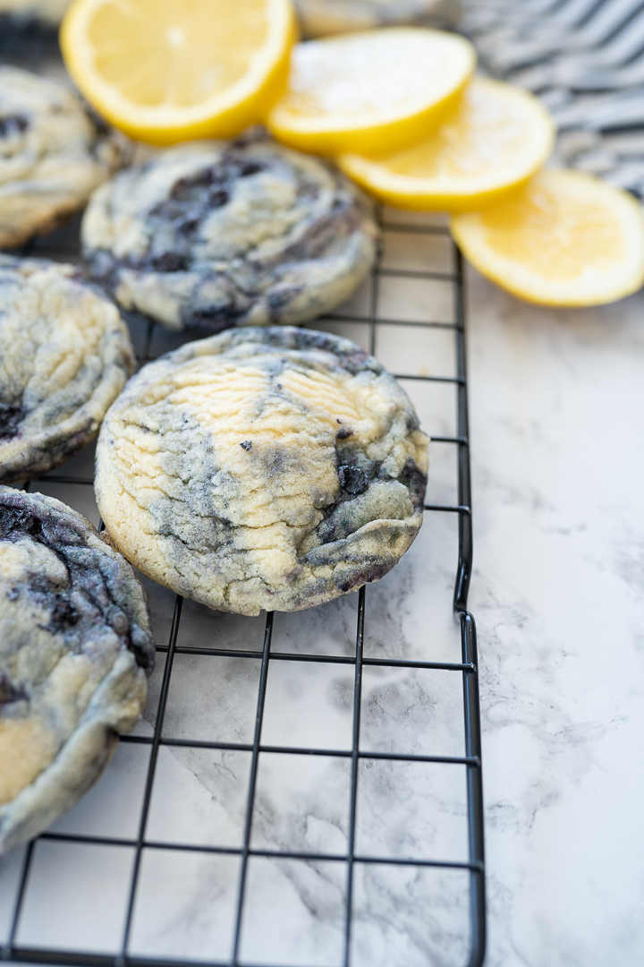 Lemon Blueberry Cookies on a cooling rack. The upper right hand corner of the cooling rack has lemon slices spilling onto the granite counter and a blue and white striped towel. The bottom right hand side of the cooling rack is empty. 