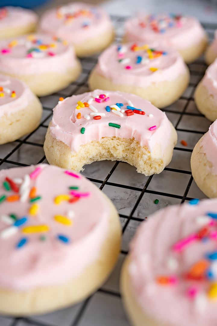 loft house sugar cookies, frosted with sprinkles on top on a cookie cooling rack. One with a bite taken out of it. 