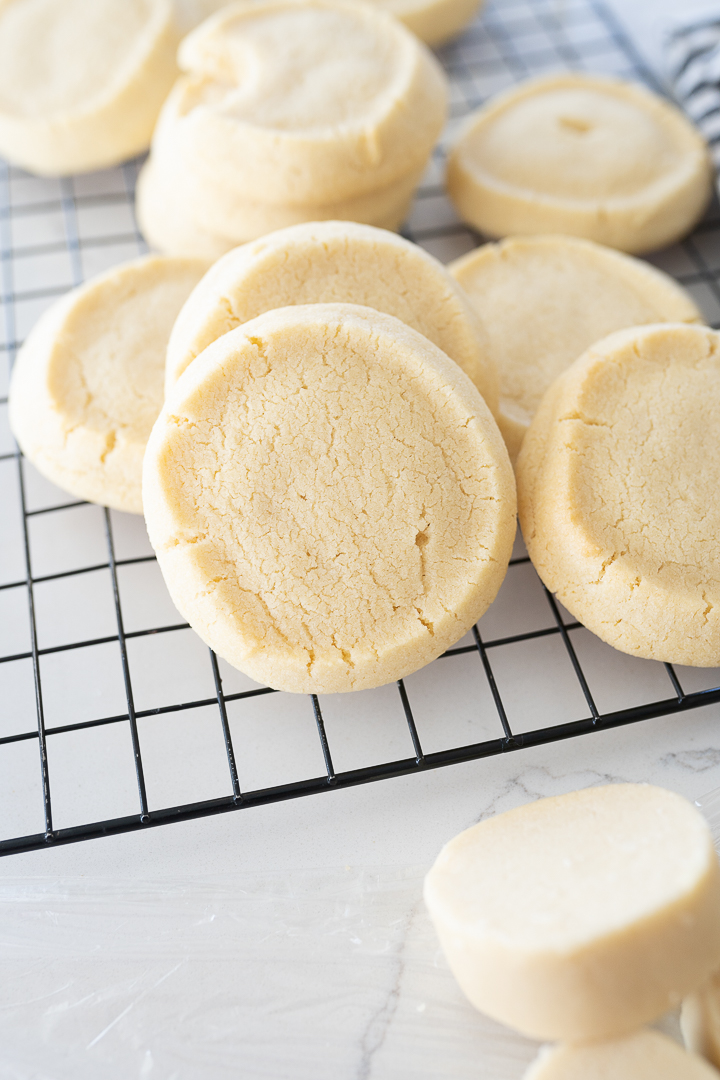 PIllsbury sugar cookies piled on a wire cooling rack. Uncooked dough in the bottom right of the frame on the counter. 