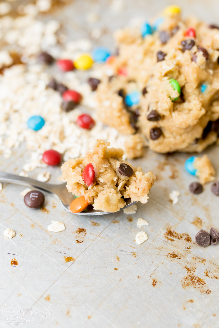 Monster cookie dough with heat-treat flour on a spoon. Behind the spoon is a pile of more cookie dough and a scattering of oats and mini m&ms. 