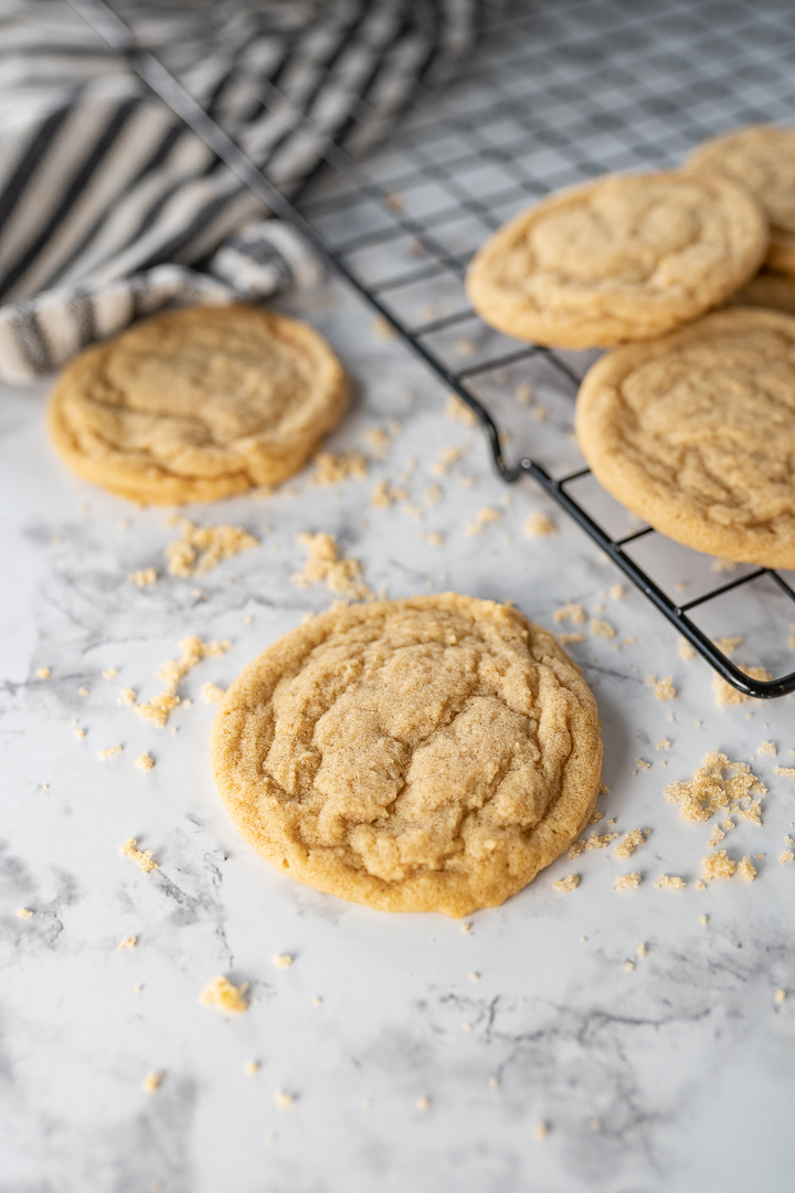 A brown sugar cookie on a marble countertop. Brown sugar is sprinkled on the counter. Next to the cookie are more brown sugar cookies on a wire cooling rack.