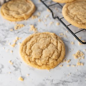 A brown sugar cookie on a marble countertop. Brown sugar is sprinkled on the counter. Next to the cookie are more brown sugar cookies on a wire cooling rack.
