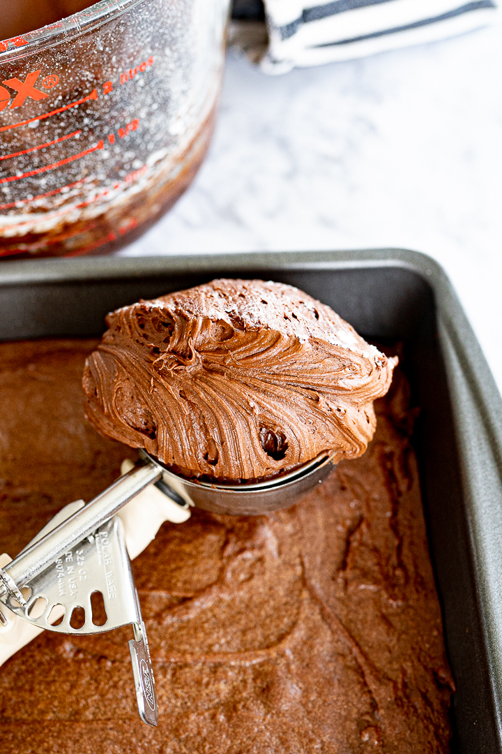 A cookie scoop filled with brownie frosting sits on top of a brownie pan. 