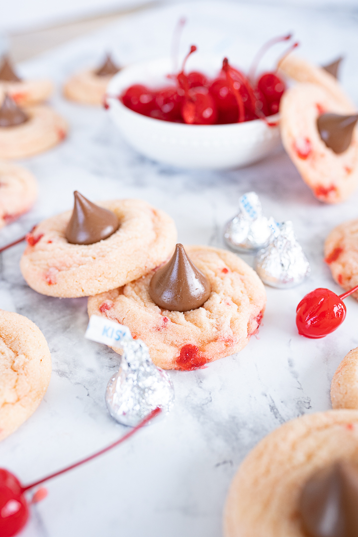 A counter full of cookies, a bowl of maraschino cherries and unopened hershey kisses. The image is focused on one cookie that is propped up on another. 