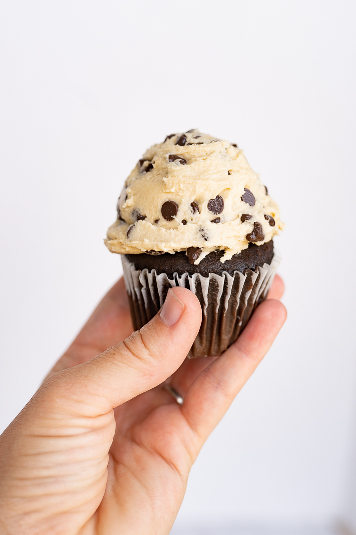 Cookie dough frosting made with heat-treat flour on top of a chocolate cupcake. The cupcake is being held up by a hand against a white wall. 