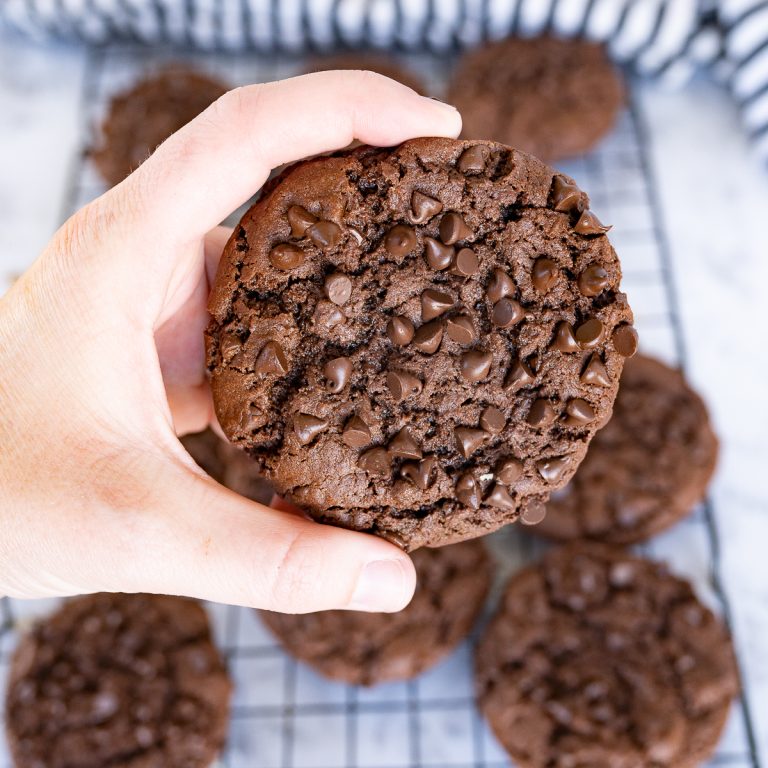 A hand holding a chocolate fudge cookie over a wire cooling rack of additional cookies.