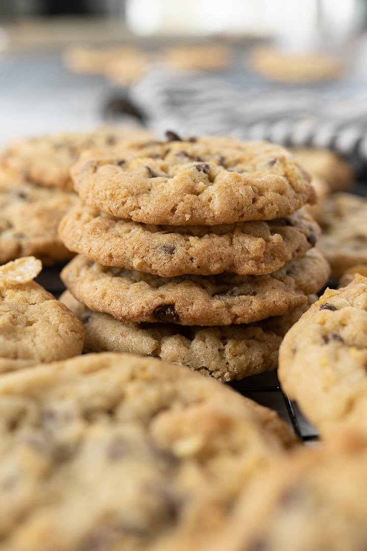 A stack of 4 cornflake cookies is in focus. Surrounding the pile of cookies are additional out of focus stacks of cookies.