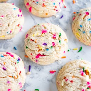 6 sprinkle birthday cake cookies on a counter top with sprinkles scattered around.