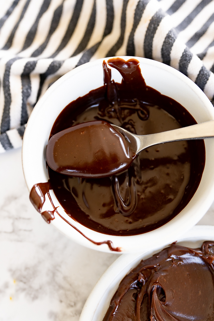 Aerial view of a bowl of chocolate ganache as it is being stirred and is still runny. The bowl is sitting on the counter next to a striped towel with a spoon of ganache being pulled out of the bowl. 