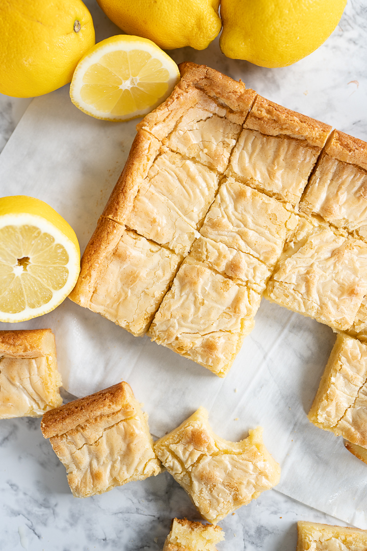A pan of lemon brownies cut into squares sits on the counter with whole lemons surrounding it. A few of the squares have been pulled away from the rest.