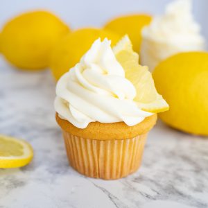 A cupcake with lemon cream cheese frosting piped on top. In the frosting is a lemon slice. In the background you can see full lemons.