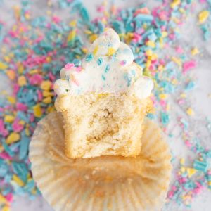 Aerial view of a cupcake that has been sliced in half laying face up on the counter top. The cupcake is topped with rainbow chip frosting and surrounded by sprinkles.