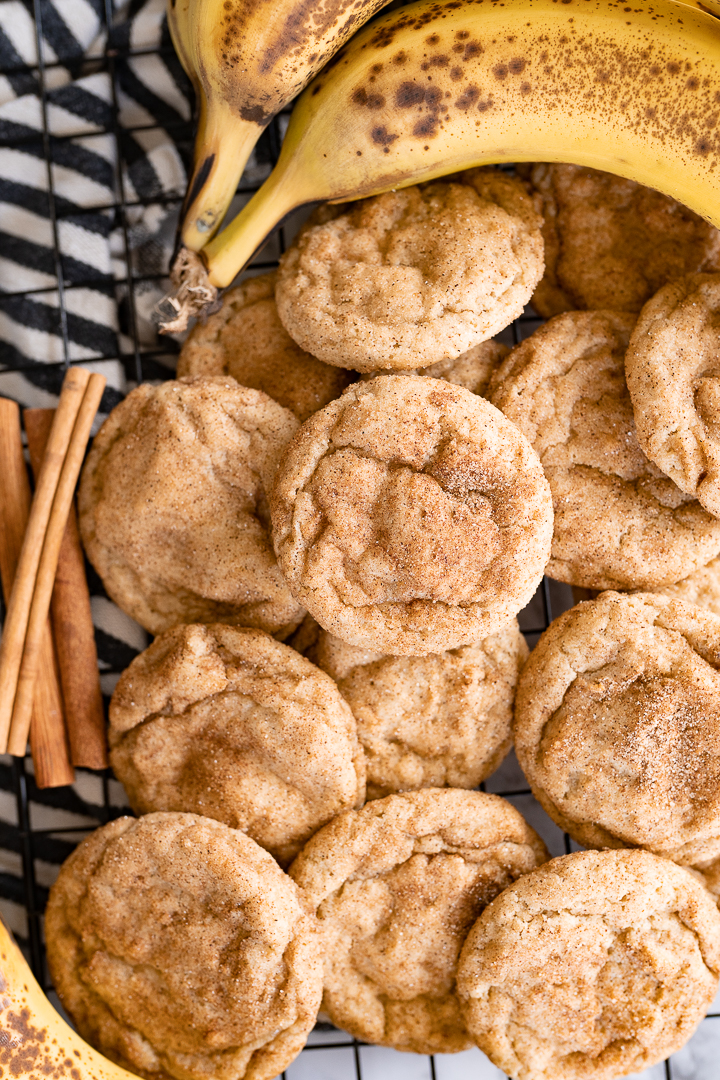 A counter filled with banana cookies rolled in cinnamon sugar. On the pile is  a stick of cinnamon and a bunch of bananas. 