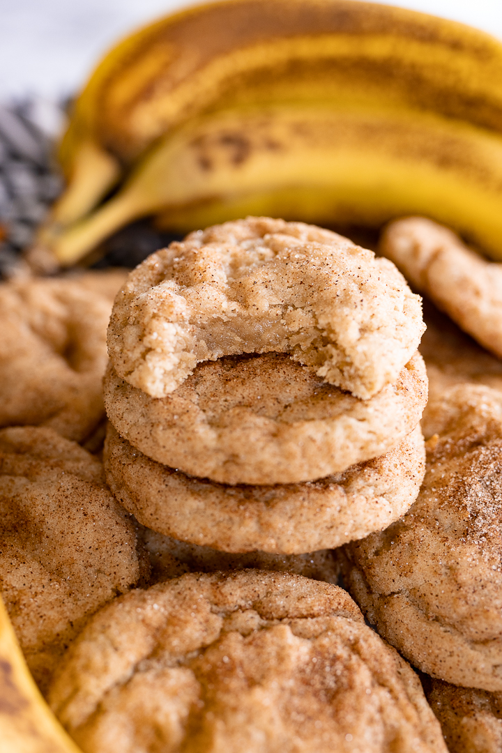A stack of banana cookies. The top one has a bite out of it. On the counter behind it is a bunch of bananas. 
