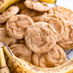 A pile of banana cookies rolled in cinnamon sugar on a counter. Next to the cookies are a few bananas.