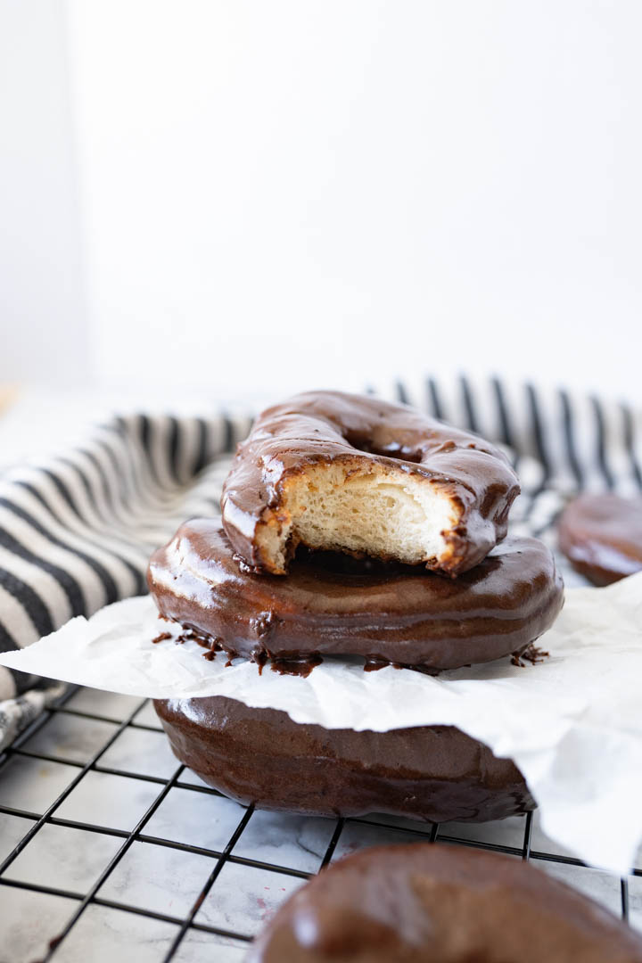 A stack of chocolate glazed donuts stacked on a cooling rack with parchment paper in between them. 