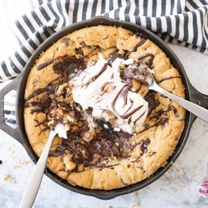 A pizookie in a skillet, topped with melty ice cream and chocolate. Two spoons stick out of the pan.