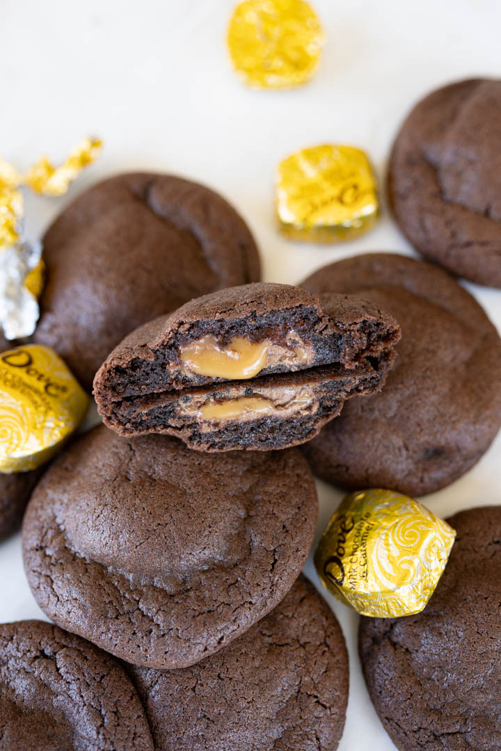 A chocolate caramel cookie cut in half and stacked back to back with the caramel oozing out. Other in-tact cookies are spread on the counter along with a dove caramel candy.