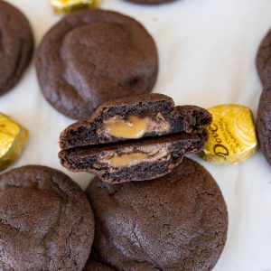 A chocolate caramel cookie cut in half and stacked back to back with the caramel oozing out. Other in-tact cookies are spread on the counter along with a dove caramel candy.