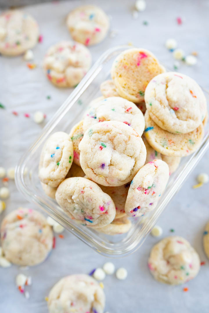 A container of white chocolate chip sprinkle cookies spilling out onto the counter. 