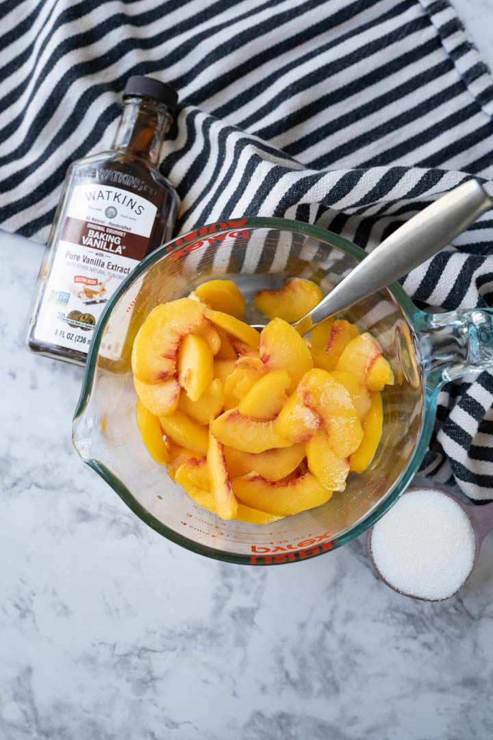 A bowl of fresh peaches sliced and ready to be made into peaches and cream. Next to the bowl is vanilla and sugar. 