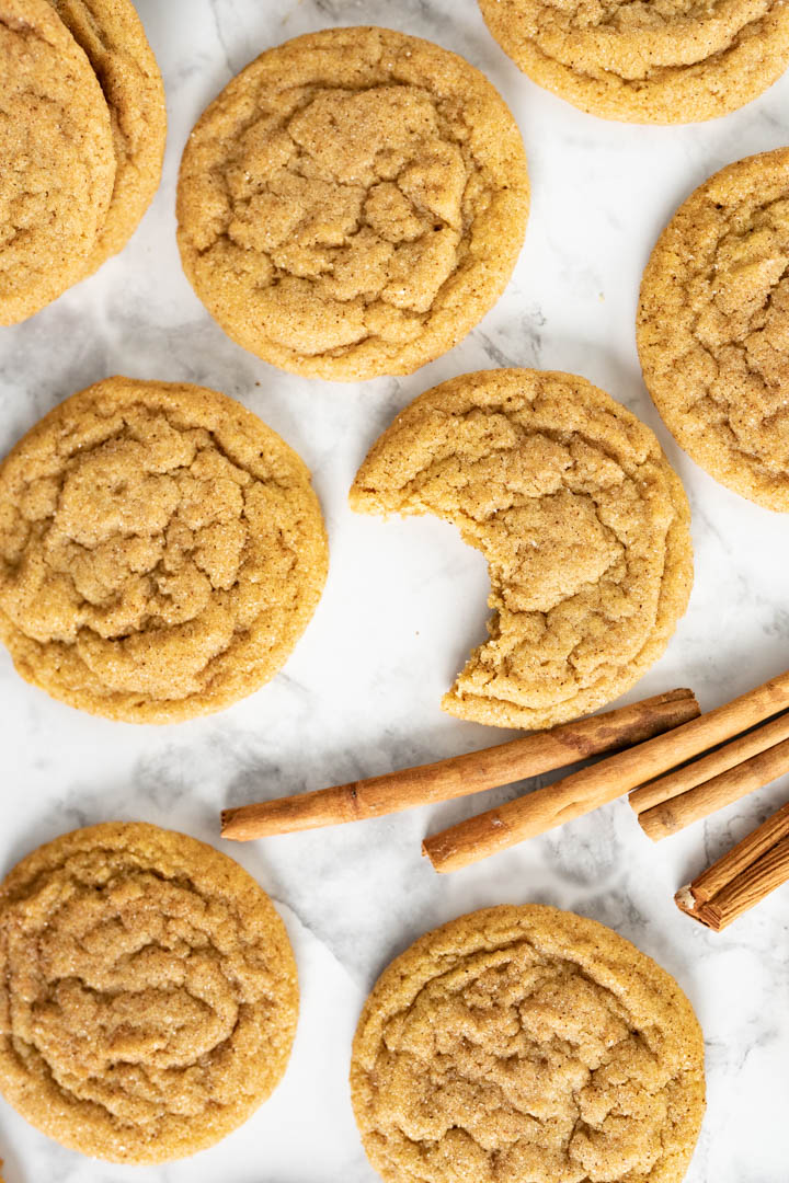 An aerial view of pumpkin cookies spread out on the counter with cinnamon sticks. One cookie has a bite out of it. 