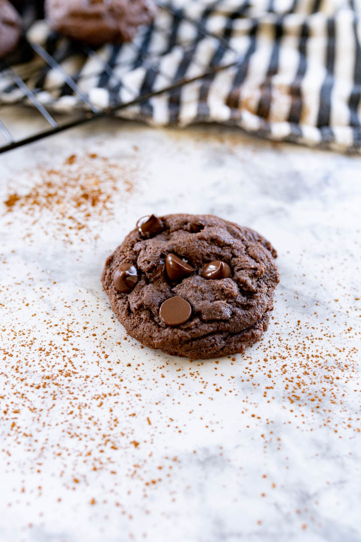 A single chocolate chocolate chip cookie on the counter surrounded by cocoa powder sprinkled around it. 