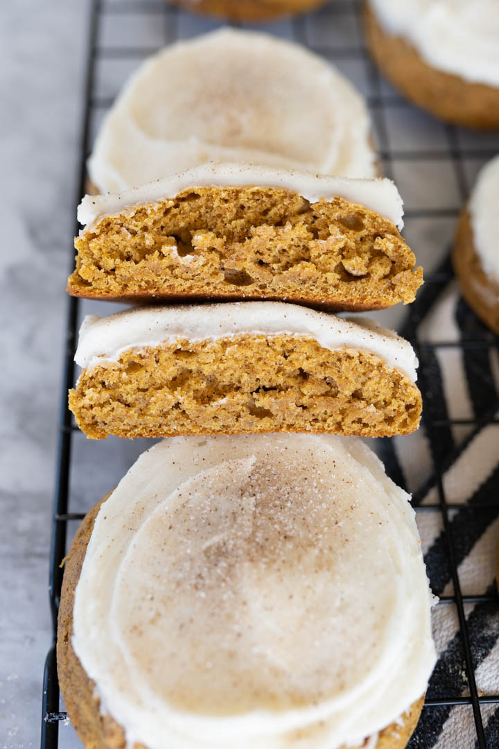 A pumpkin cookie with cream cheese frosting cut in half stacked together , leaning on another cookie. 