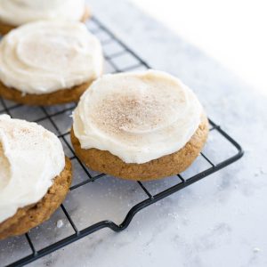 Angled image of 3 pumpkin cookies with cream cheese frosting on the corner of a wire cooling rack.